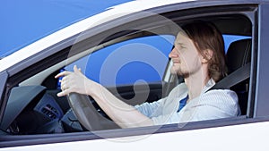 Happy funny man is listening to music and dancing in the car. Young driver is sitting in white car with blue background