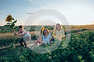 Happy funny smiling family on the picnic, serious boy in casual clothes crouched with sunflower in hands, her mom in