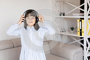 Happy funny teen girl with dark hair wear headphones standing in room and listening new pop music, enjoying, singing song,