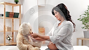 Happy future mother pregnant woman with belly playing bear soft toy waiting baby at white room