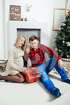 Happy future parents are sitting on the carpet in the room decorated to Christmas holidays. Pregnant woman is observing