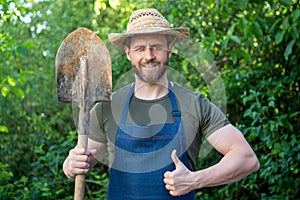 Happy gardener man in gardening apron and farmers hat with garden spade giving thumb gesture