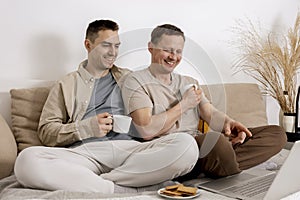 Happy gay couple with casual clothes spending time together at home, watching movie on the laptop and drinking tea. Two