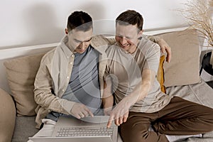 Happy gay couple with casual clothes spending time together at home and watching movie on the laptop. Two caucasian men