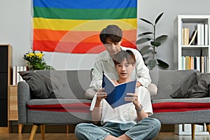 Happy gay couple reading book on sofa at home, spending time together on weekend. LGBTQ people lifestyle and love