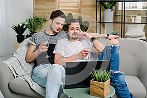 Happy gay couple sitting on sofa, watching movie and drinking wine. Gay couple, homosexual concept