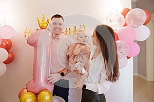 Happy and gentle parents hold baby daughter with birthday balloon decor.