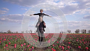 Happy Germans family, young male with child boy spread arms to side sitting on shoulders walk across floret field of