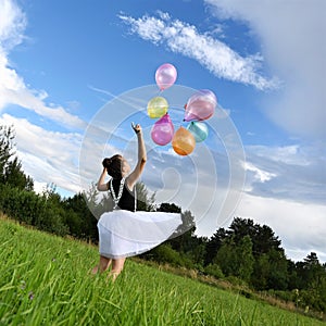 happy girl beautiful dress is holding colorful balloons in summer meadow against background of blue sky and clouds