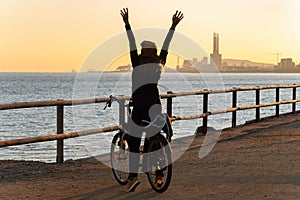 Happy girl with a bicycle raised her hands up and enjoys the sunset on the promenade road of Maresme, Catalonia, Spain photo