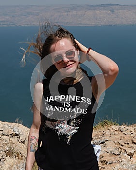 a happy girl in a black T-shirt and sunglasses stands against the backdrop of a mountain landscape with a pond