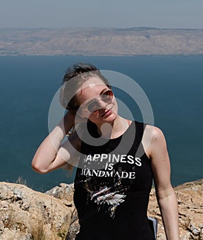 A happy girl in a black T-shirt and sunglasses stands against the backdrop of a mountain landscape with a pond