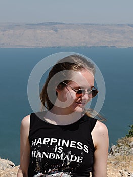 A happy girl in a black T-shirt and sunglasses stands against the backdrop of a mountain landscape with a pond