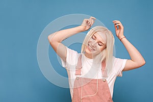 Happy girl blond girl listens to music in headphones with eyes closed on a blue background, dancing and smiling. Smiling teen girl