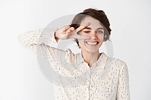 Happy girl in blouse showing peace or victory sign on eye and winking, express positivity and joy, standing against