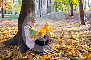 Happy girl with a bouquet of maple yellow autumn leaves sitting under a tree in a sunny city park. Life style