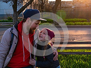 Happy girl with braided hair and father in spring park sits on a bench on sunset outdoor.Kid with dad smiling having fun