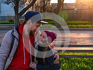 Happy girl with braided hair and father in spring park sits on a bench on sunset outdoor.Kid with dad smiling having fun