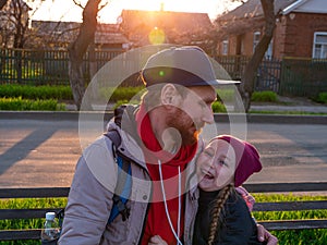 Happy girl with braided hair and father in spring park sits on a bench on sunset outdoor.Kid with dad smiling having fun