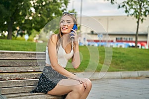 Happy girl calling on cell phone looking away sitting on a bench in the street. Young blonde girl waiting for a person