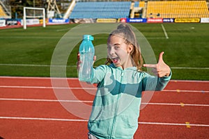 Happy girl child in activewear stand at sports stadium pointing finger at sport water bottle, hydration