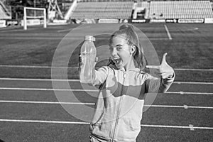 Happy girl child in activewear stand at sports stadium pointing finger at sport water bottle, hydration