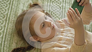 Happy girl child with blond hair and pigtails, lying on the sofa, using the phone, smiling, portrait