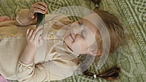 Happy girl child with blond hair and pigtails, lying on the sofa, using the phone, smiling, portrait