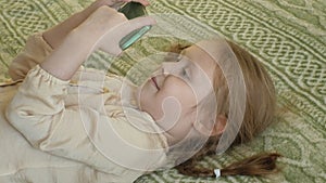Happy girl child with blond hair and pigtails, lying on the sofa, using the phone, smiling, portrait