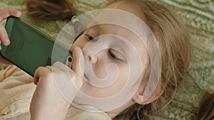 Happy girl child with blond hair and pigtails, lying on the sofa, using the phone, smiling, portrait