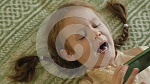 Happy girl child with blond hair and pigtails, lying on the sofa, using the phone, smiling, portrait