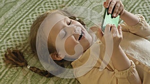 Happy girl child with blond hair and pigtails, lying on the sofa, using the phone, smiling, portrait