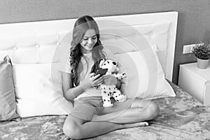 Happy girl child with plush stuffed puppy toy use mobile phone sitting on bed, sms