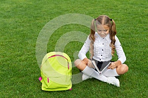 Happy girl child with school bag reading book sitting on green grass