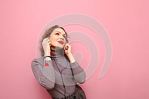 Happy girl with curly hair listens to music in headphones with a smile on her face, looks up at copy space, isolated on pink