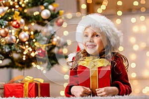 The happy girl is delighted with the gift and looks into the distance. A child next to a decorated Christmas tree