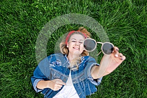 Happy girl in a denim jacket lying on a green lawn and holding sunglasses in her hands