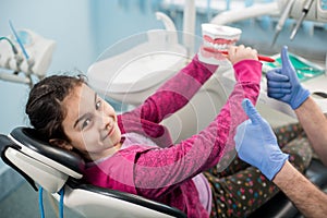 Happy girl in dentist chair educating about proper tooth-brushing in dental clinic. Dentistry, oral hygiene concept