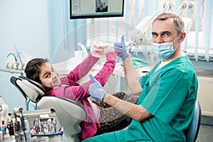 Happy girl in dentist chair educating proper tooth-brushing, using dental jaw model and toothbrush in dental office