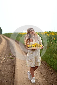 a happy girl on a dirt unpaved road going into the distance holds a wooden box with sunflower flowers