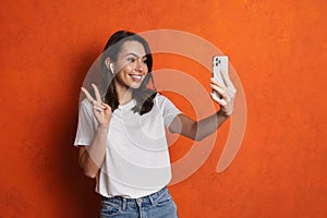 Happy girl in earphones gesturing peace sign while taking selfie