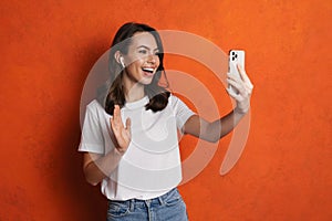 Happy girl in earphones waving hand while taking selfie on cellphone
