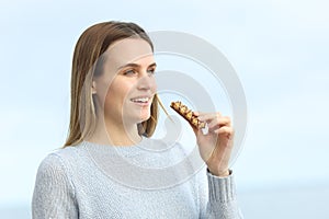 Happy girl eating cereal bar looking away on the beach