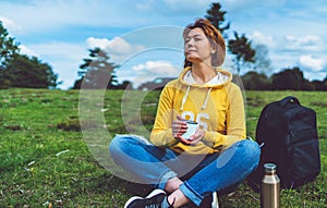 Happy girl with eyes closed holding in hands cup of hot tea on green grass in outdoors nature park, beautiful woman hipster enjoy