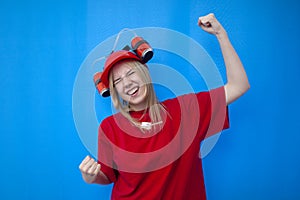 Happy girl fan in red uniform rejoices victory on a blue background, a cheerleader with a beer hat shows victory gesture