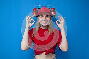 Happy girl fan in red uniform shows ok gesture and smiles on a blue background, funny cheerleader with beer hat