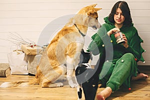 Happy girl in festive pajamas feeding dog and cat with canned food, sitting on floor with christmas presents in stylish room.