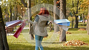 Happy girl with foxy hair spinning in park with purchases in multi-colored paper bags and rejoices