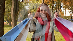 Happy girl with foxy hair walks in the park with purchases in multi-colored paper bags and rejoices