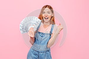 Happy girl holding cash dollar bills and showing a winning gesture on an isolated pink background